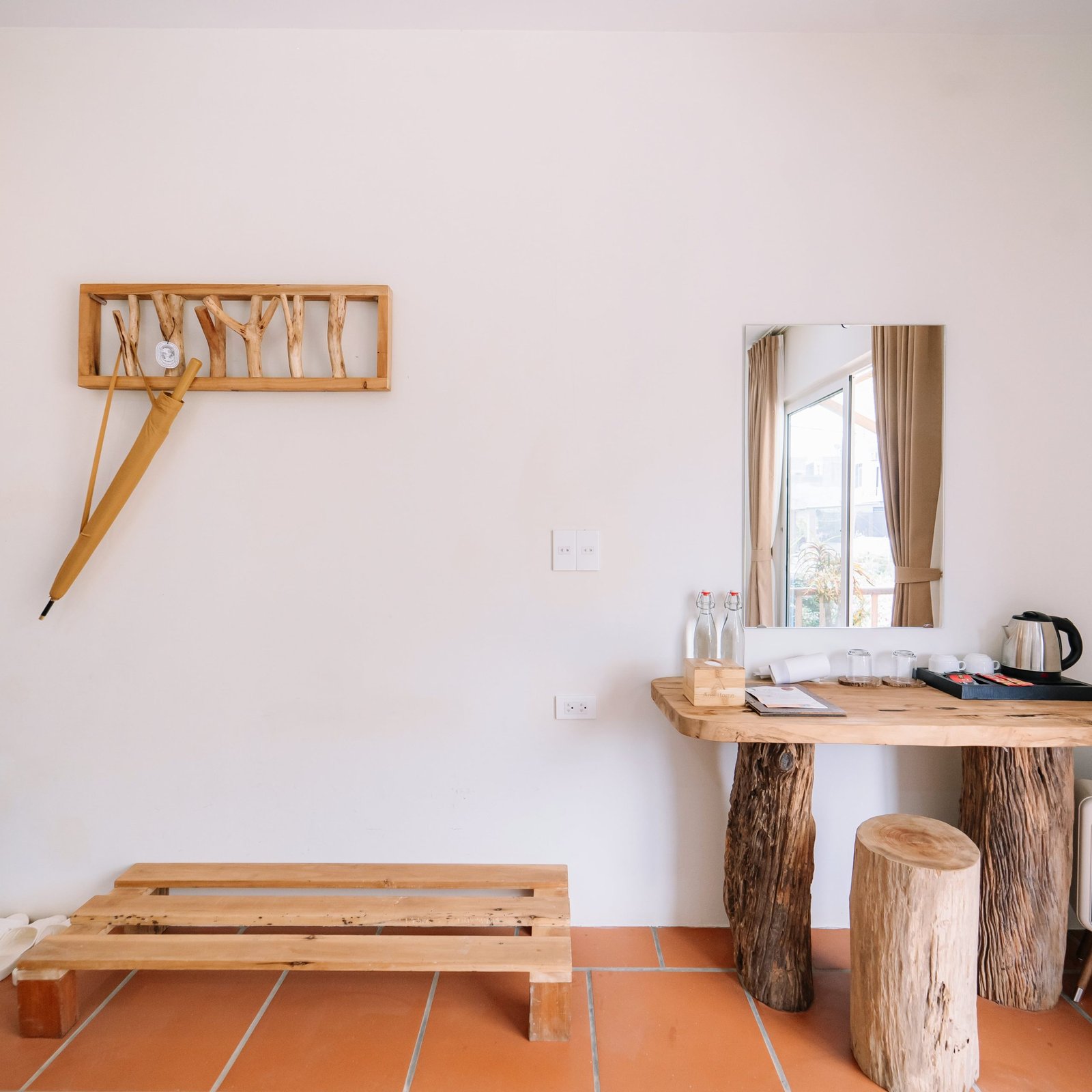 A timber console with a kettle and glasses, a mirror and a branch coat rack on the wall